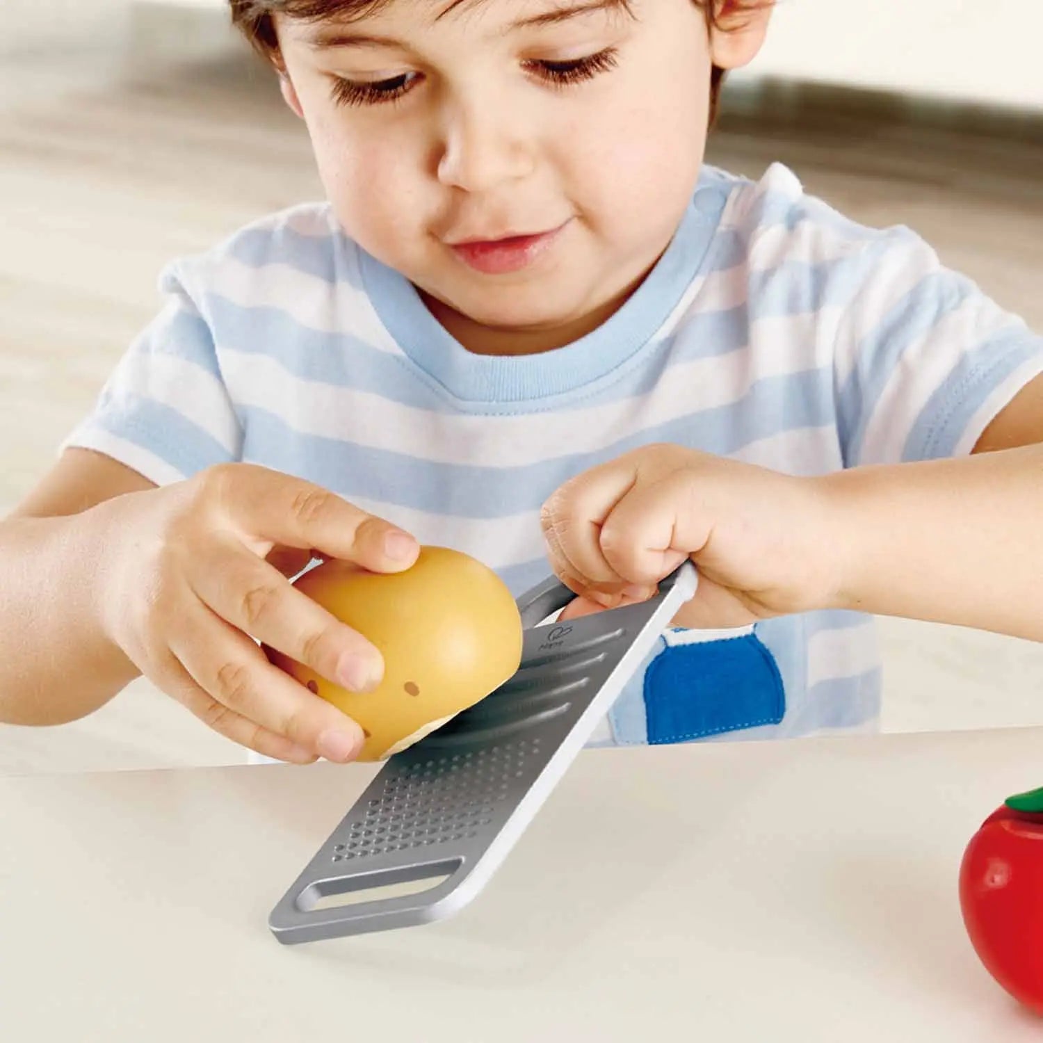A young boy focuses on grating a yellow toy fruit with a toy grater.