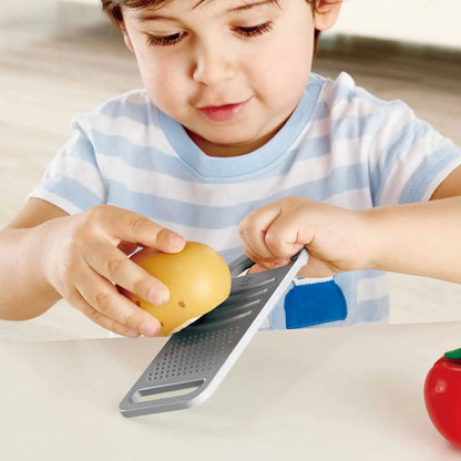 A young boy focuses on grating a yellow toy fruit with a toy grater.