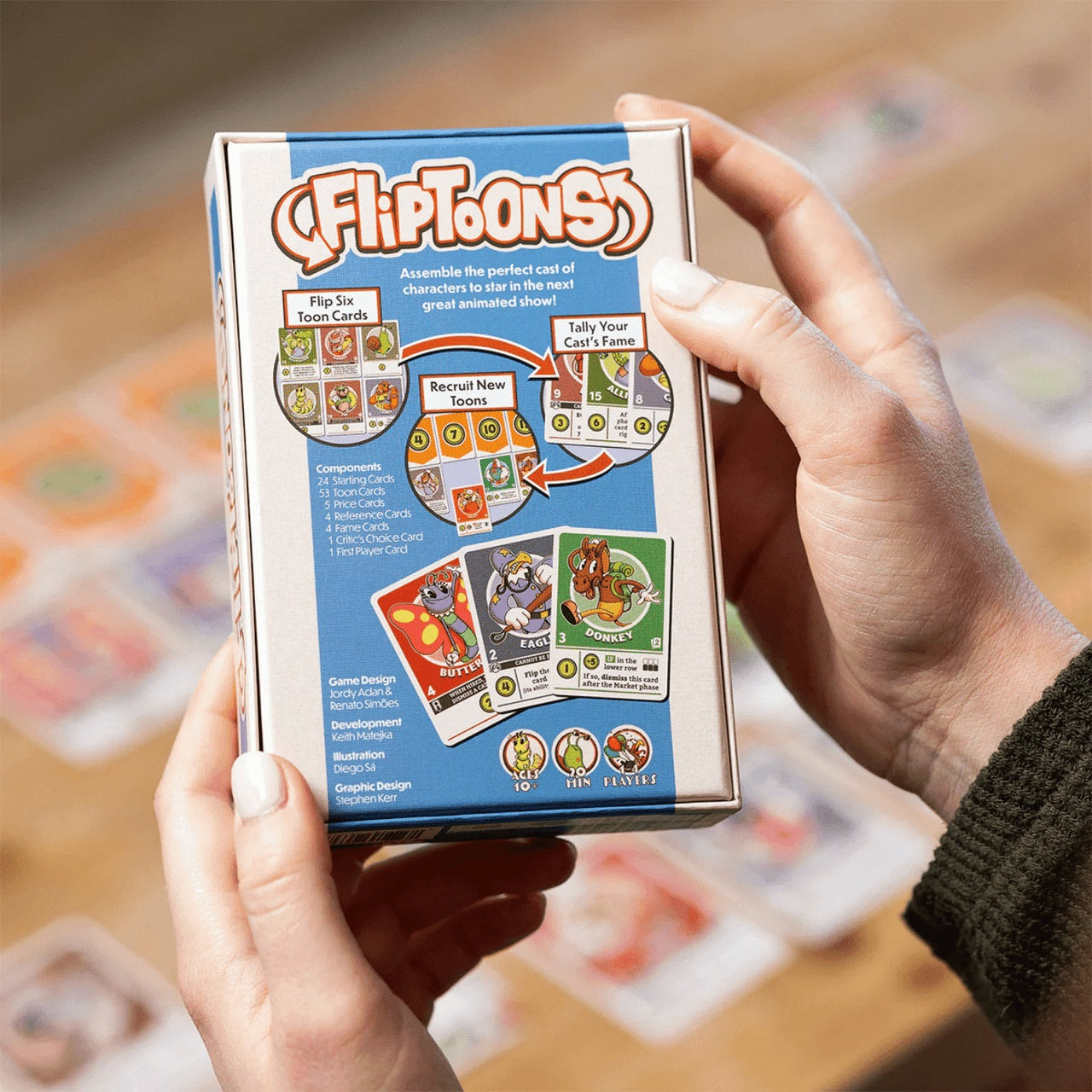 Hands holding the back of the FLIPTOONS card game box, featuring rules and cartoon animal artwork