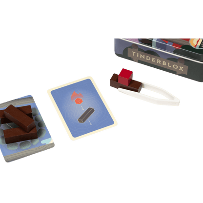 Tinderblox game set: wood blocks, challenge cards, white tongs holding a red block, and the game tin