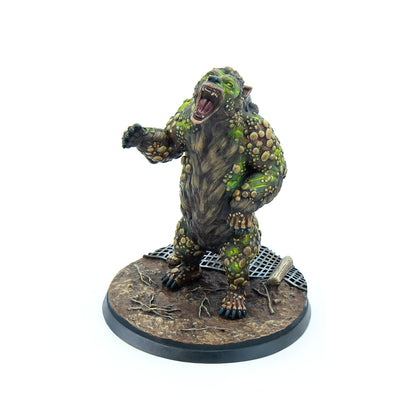A miniature monstrous creature with brown fur and green, bumpy growths, snarling with an open mouth and extended arm, on a gritty base