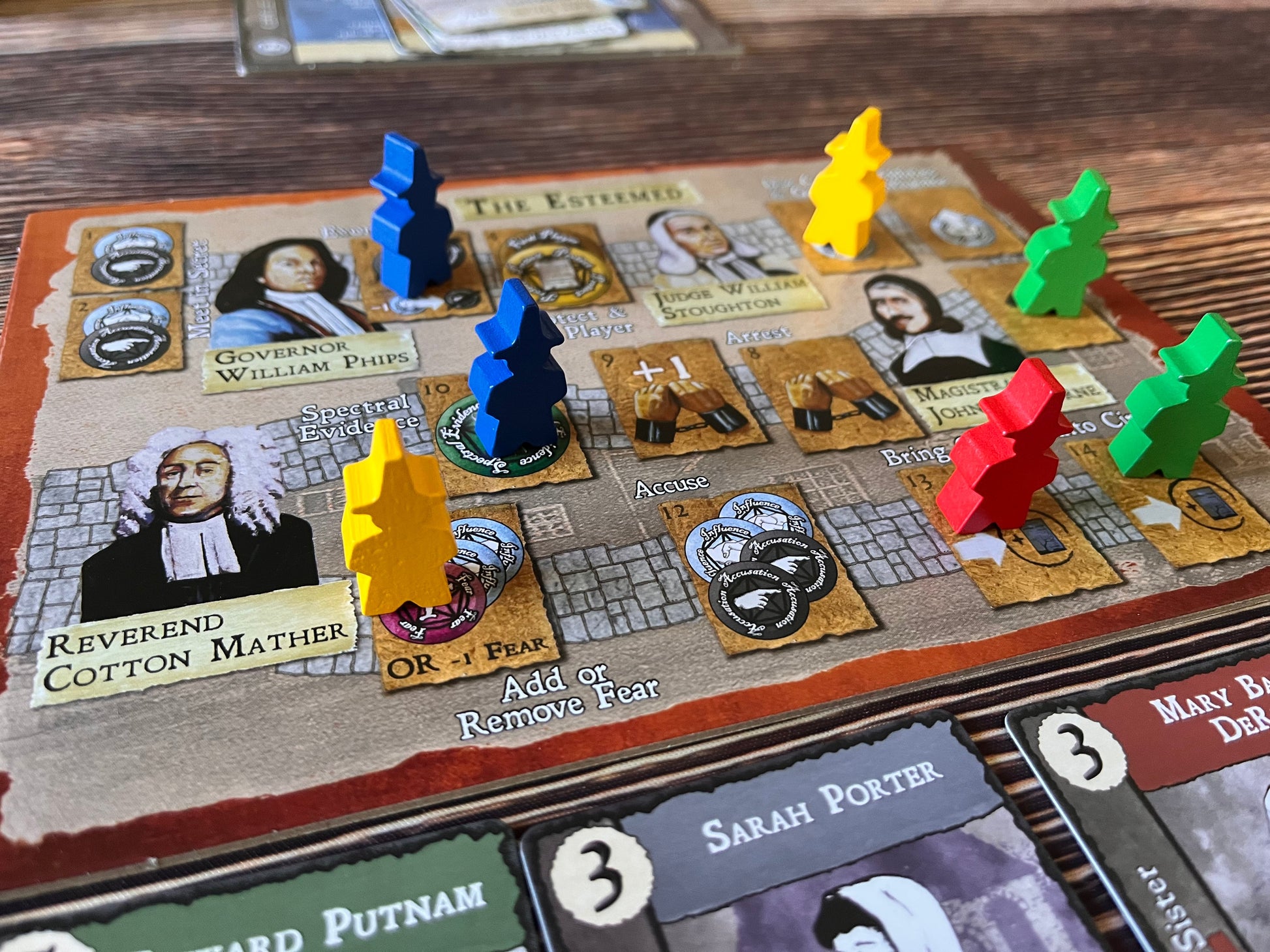 A close-up of a board game with blue, yellow, red, and green pawns on action spaces like 'Accuse' and 'Spectral Evidence'