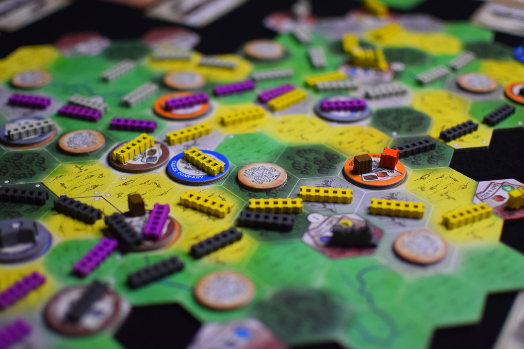 A colorful board game with a hexagonal map. Green and yellow terrain tiles are covered with wooden blocks and round tokens