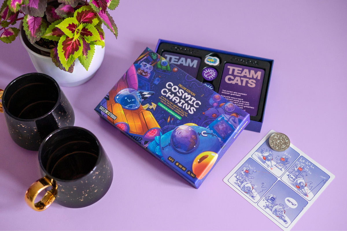 A Cosmic Chains Deluxe Edition board game box, an open &quot;Team Cats&quot; game, two black mugs, a plant, and a comic card on a purple surface