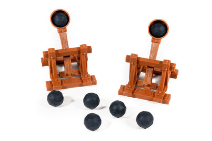 Two brown toy catapults, each loaded with a black ball, with four additional black balls scattered on a white surface