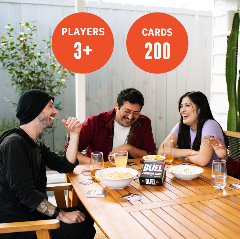 Three laughing adults play &quot;Duel: A Drinking Game&quot; at an outdoor table with snacks and drinks