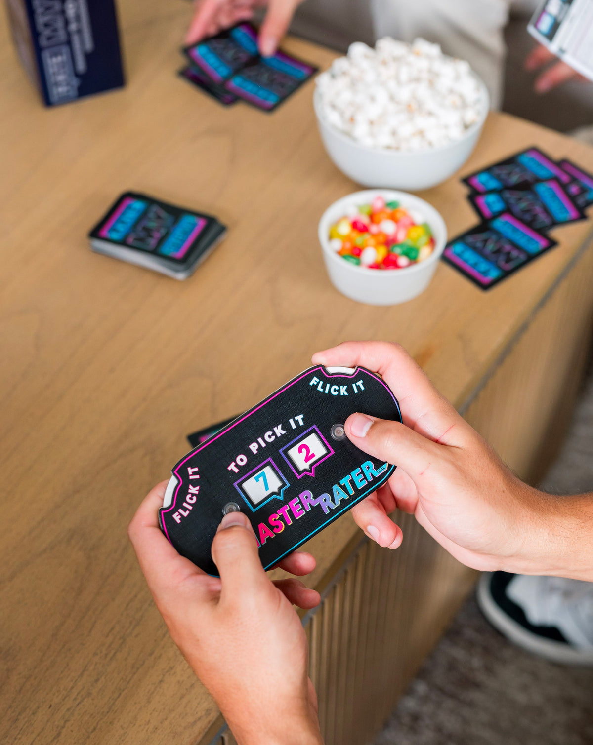 Hands hold a black Master Rater card game with a 7 and 2 displayed surrounded by game cards and snacks on a table