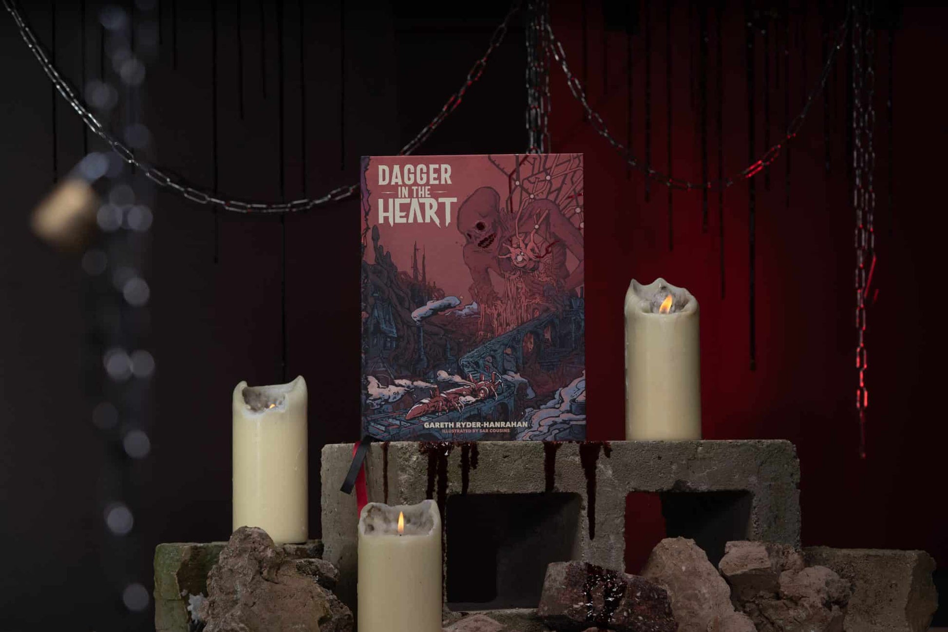 Gothic display of the book "Dagger in the Heart" surrounded by three candles, set atop concrete blocks against a red backdrop