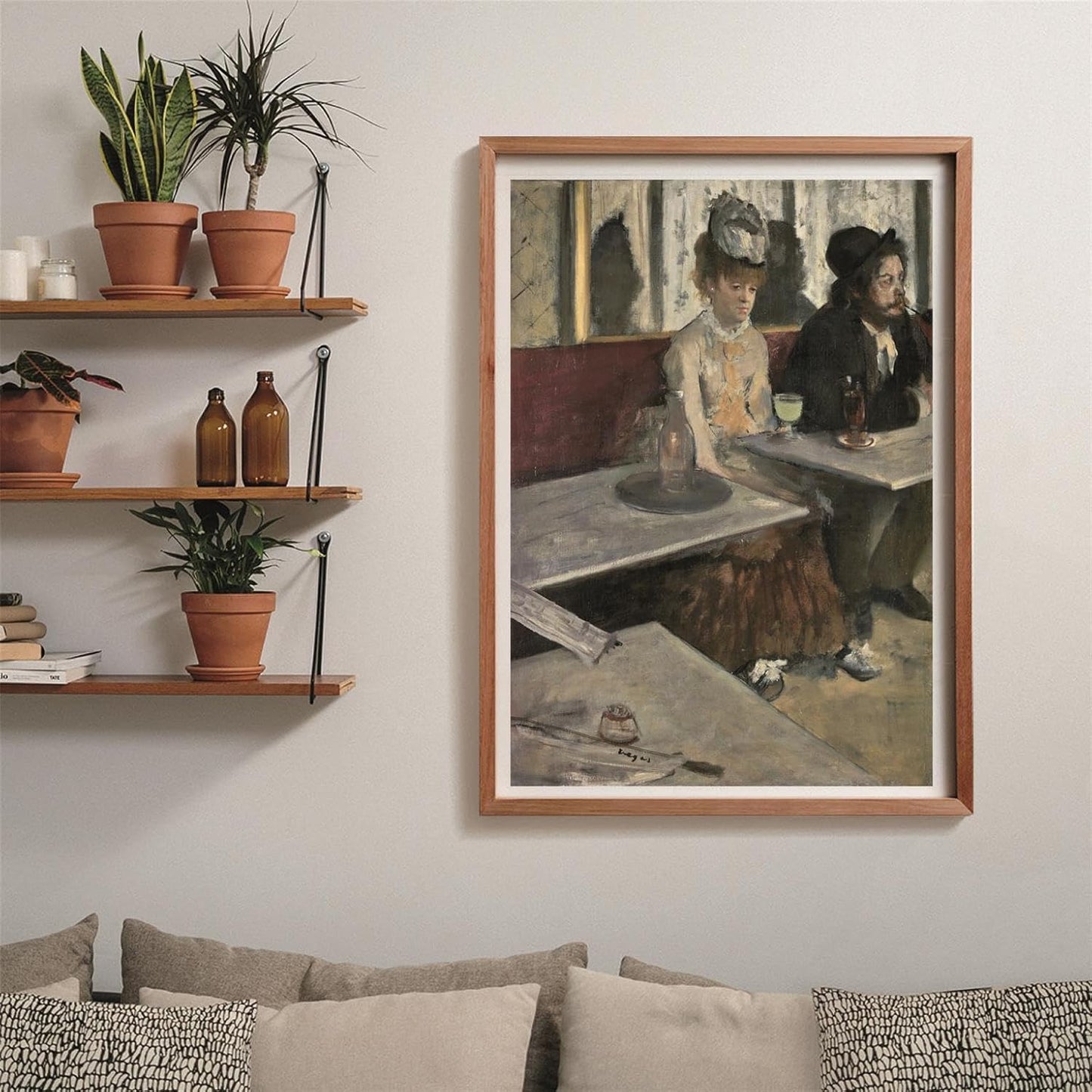 A framed painting of two somber people in a cafe hangs on a light wall, with potted plants on shelves and a sofa below