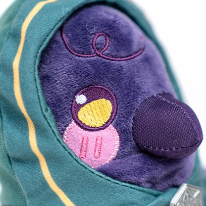Close-up of a fluffy purple plush toy with one large yellow and white embroidered eye, pink lower face, and dark purple nose, wearing a striped teal hood
