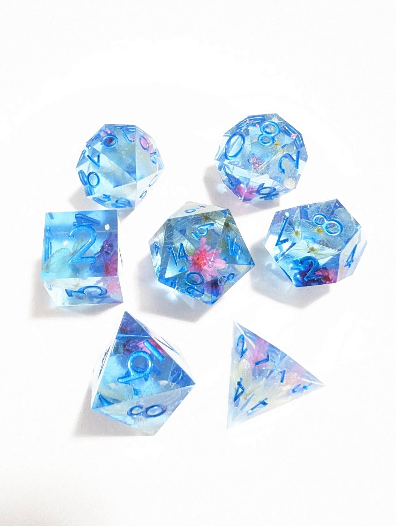 Seven translucent light blue polyhedral dice, embedded with pink and purple flowers, display blue numbers on a white surface