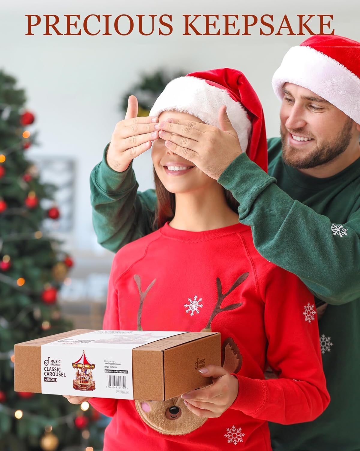 Man surprises woman with a &quot;Classic Carousel Kit&quot; gift while covering her eyes. Both wear Santa hats