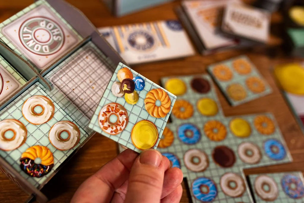 Donut Shop board game from 25th Century Games showcasing various donut tiles including a hand holding a tile with assorted donut art