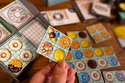 Donut Shop board game from 25th Century Games showcasing various donut tiles including a hand holding a tile with assorted donut art