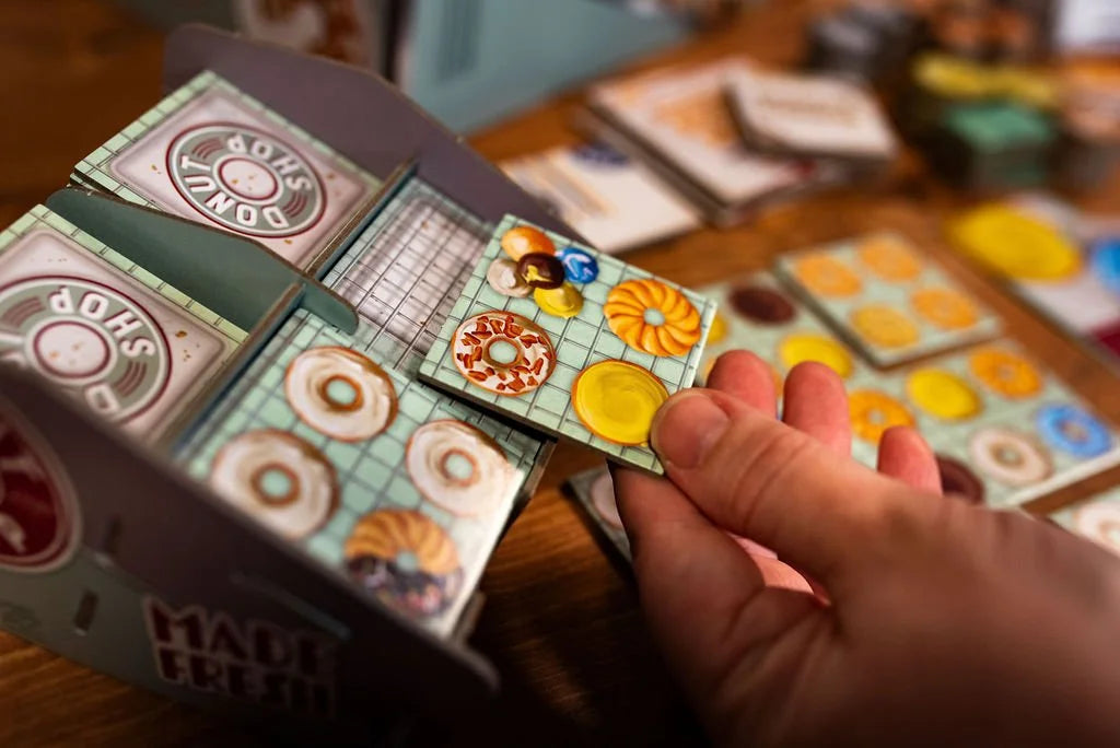 25th Century Games Donut Shop board game components including a hand holding a game tile with four colorful donuts beside stacked Donut Shop game boxes