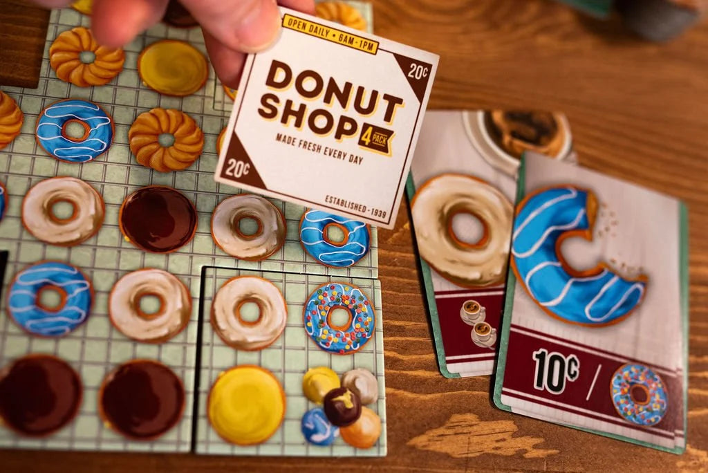 25th Century Games Donut Shop board game with player&#39;s hand holding a Donut Shop card showcasing various donut tiles and player cards including a blue frosted donut with sprinkles and a partially eaten blue donut card