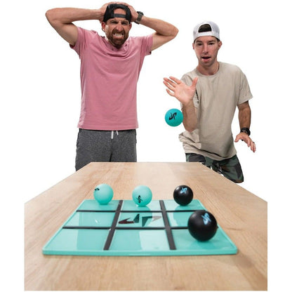 Two men play a ball-toss game on a tic-tac-toe board; one throws a turquoise ball, the other, frustrated, holds his head