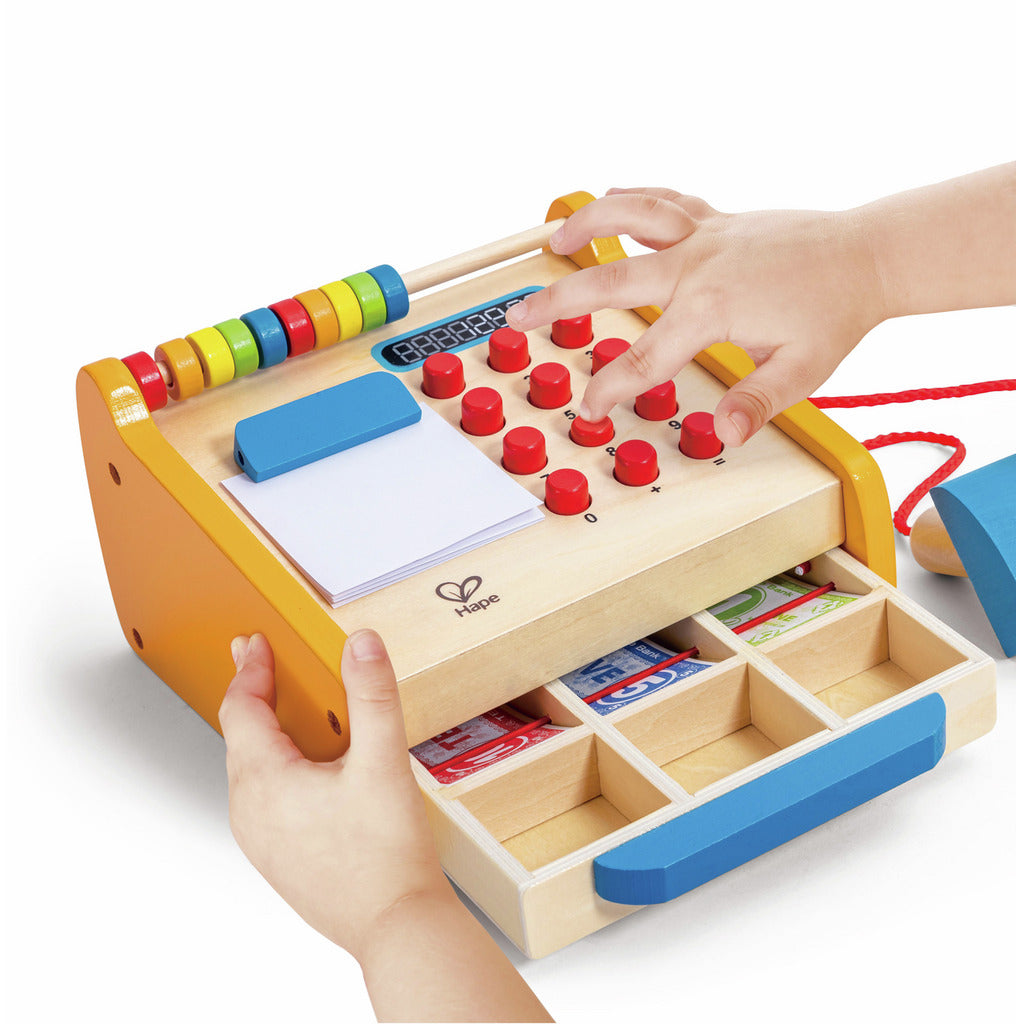 Child's hands playing with a wooden Hape cash register toy, featuring red buttons, a colourful abacus, and an open cash drawer