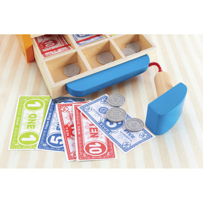 Wooden toy cash register drawer with paper bills (1, 5, 10) and grey plastic coins on a striped surface