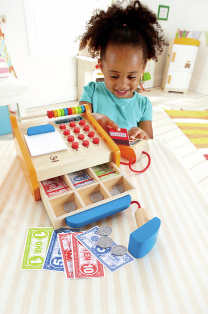 Smiling girl playing with a wooden toy cash register, swiping a card and showing play money