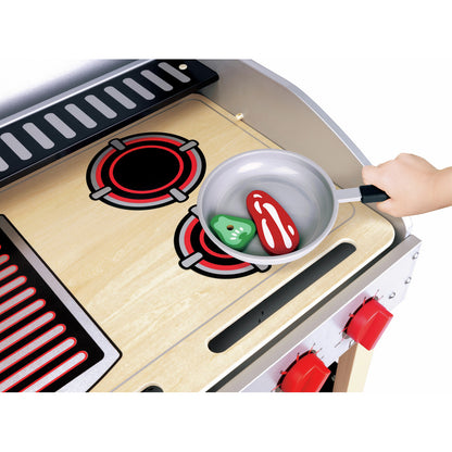 Toy stove cooktop with a pan, red and green wooden food, and a child's hand