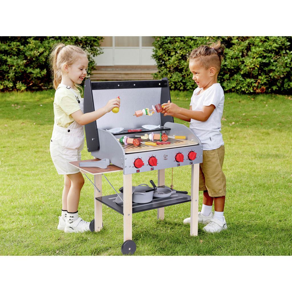 Two children grilling wooden play food skewers and sausages on a toy BBQ set outdoors