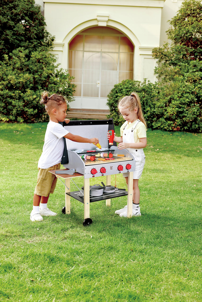 Children grilling toy food on a wooden play BBQ set outdoors in a manicured garden setting