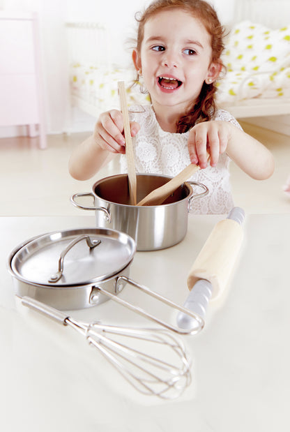 Smiling girl stirring a steel toy pot with wooden spoons, whisk and rolling pin in the foreground