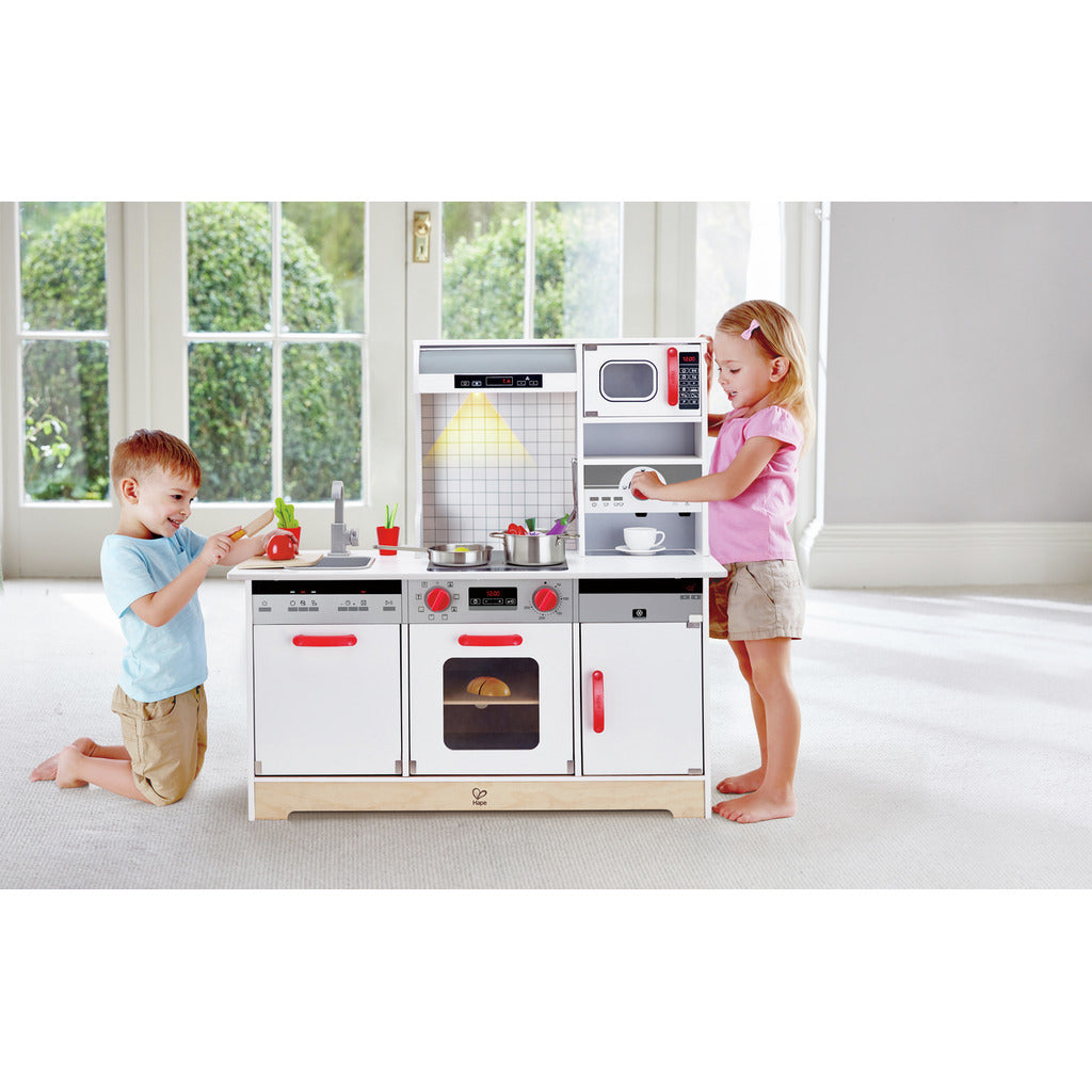 Two children playing with a modern Hape wooden play kitchen, featuring an oven, microwave, and sink