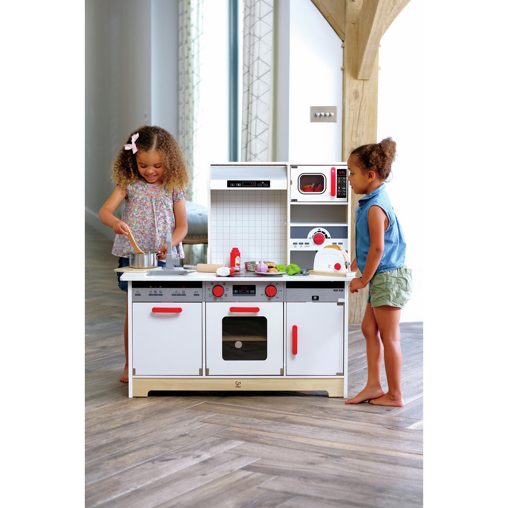 Two children playing with a modern white wooden toy kitchen set, featuring an oven, microwave, and realistic accessories