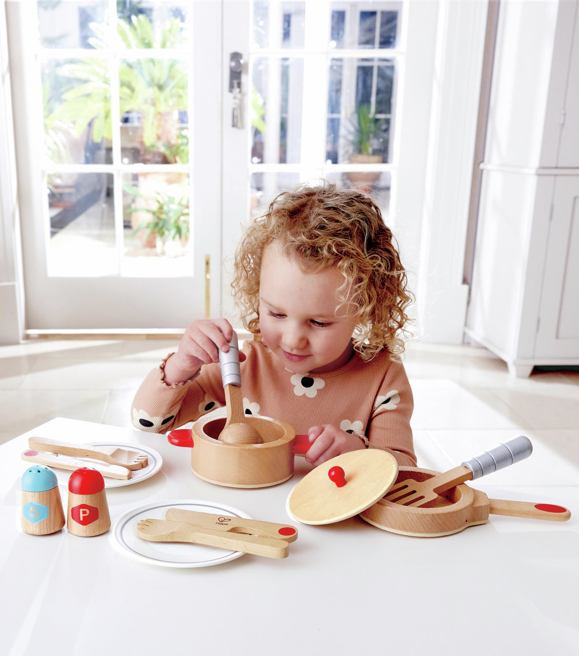 Young girl stirring a wooden toy cooking pot with a spoon, surrounded by wooden play dishes and salt and pepper shakers