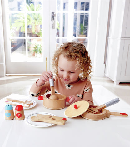 Young girl stirring a wooden toy cooking pot with a spoon, surrounded by wooden play dishes and salt and pepper shakers