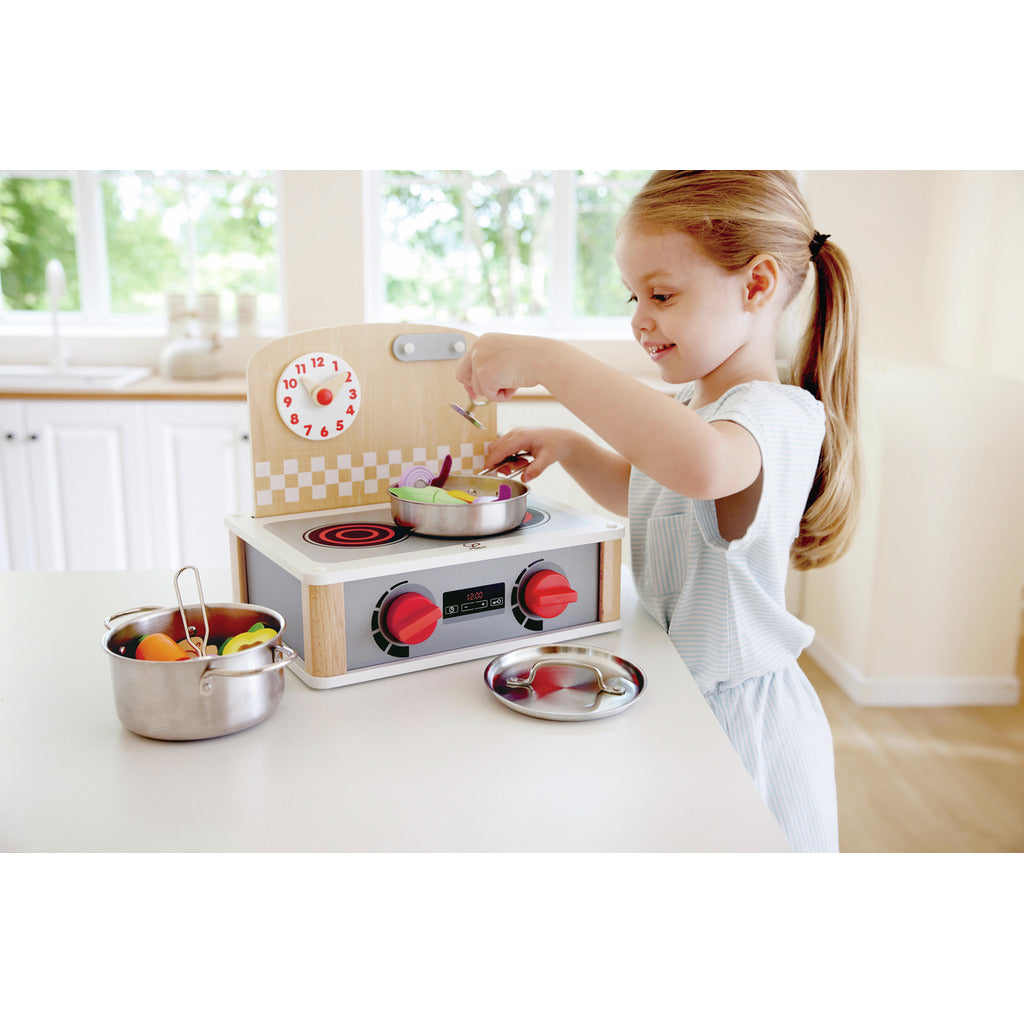Happy girl playing with a wooden toy kitchen stove, stirring colourful pretend vegetables in a stainless steel pot