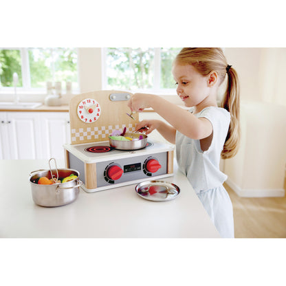 Happy girl playing with a wooden toy kitchen stove, stirring colourful pretend vegetables in a stainless steel pot