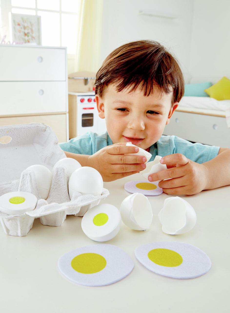 Young boy cracking a plastic toy egg. Felt yolks, egg slices, and an egg carton are on the table