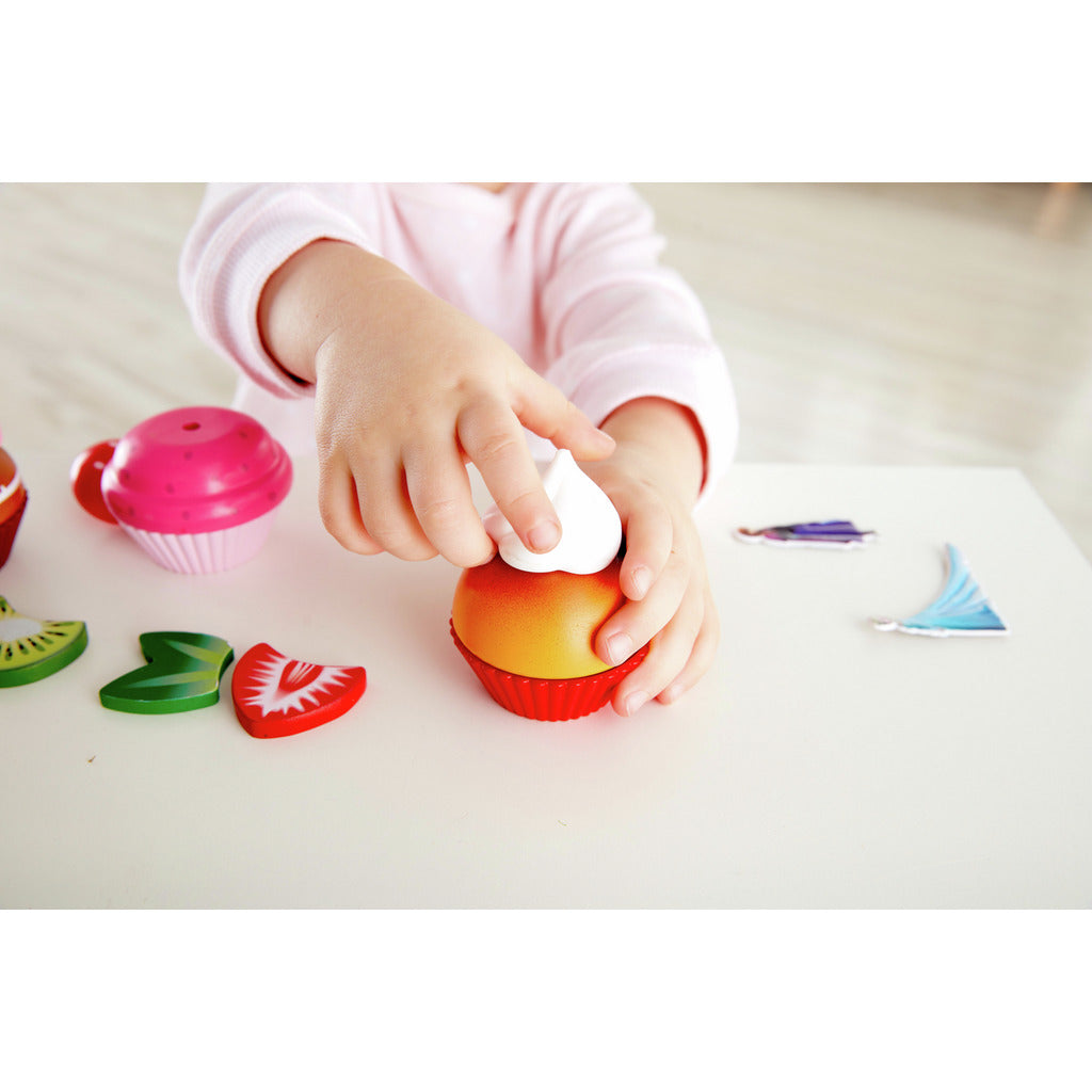 Child's hands placing toy whipped cream onto a wooden play food cupcake surrounded by fruit slices