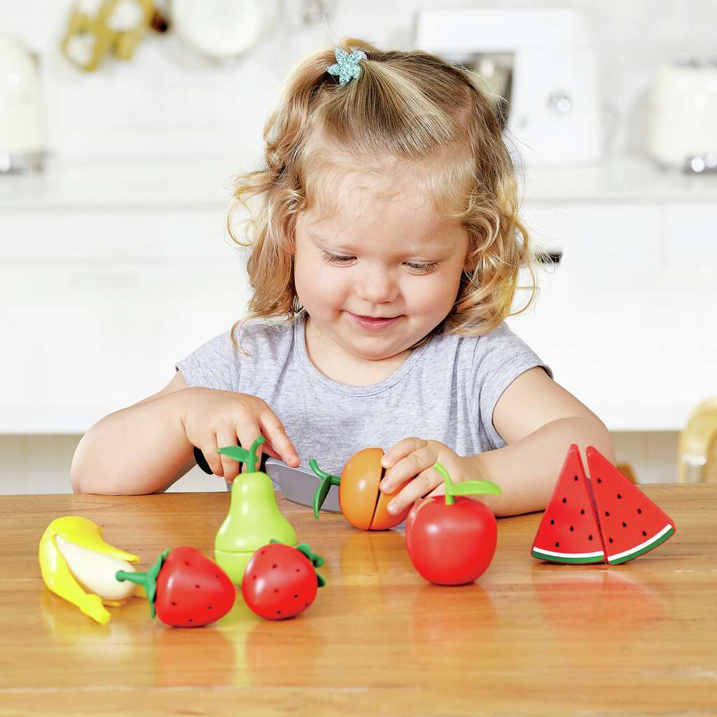 Smiling girl using a play knife to cut a toy orange, surrounded by pretend fruit