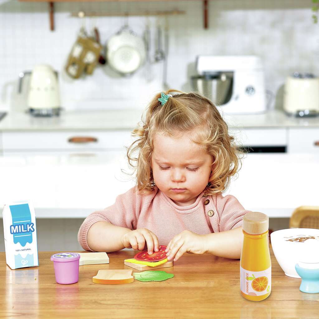 Toddler building a toy sandwich using wooden play food pieces on a kitchen table