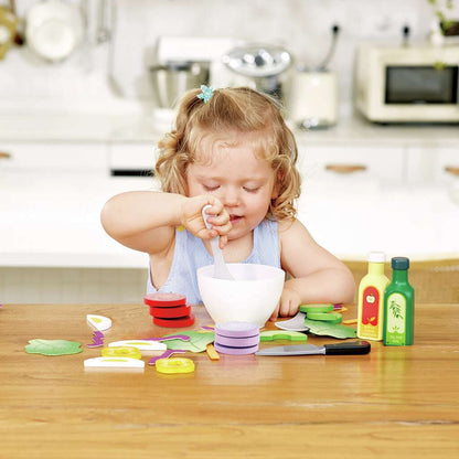 Young child stirring play food in a bowl, surrounded by toy vegetables and condiments