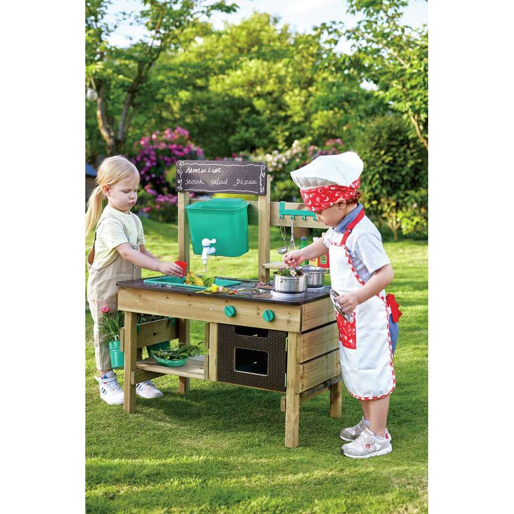 Two children play with a wooden outdoor mud kitchen on a sunny lawn, one wearing a chef hat