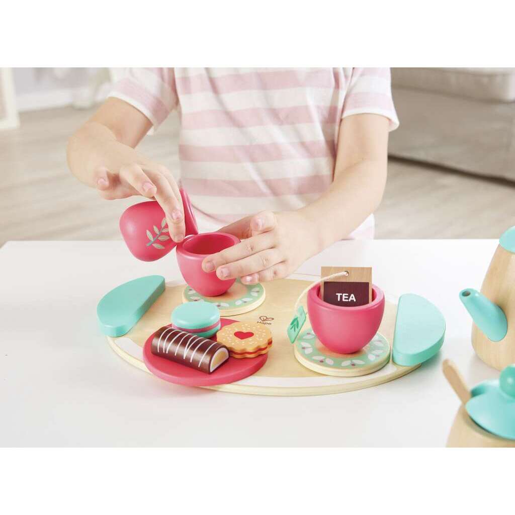 Child pouring from a pink toy pitcher into a teacup on a wooden tea set tray with desserts