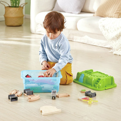 Toddler kneeling on a wooden floor, pulling wooden train tracks from a blue storage container