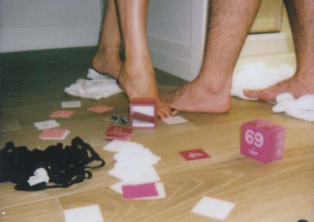 69 naughty game for couples box in pink with the number 69 visible surrounded by scattered white and pink cards on a light wood floor with two pairs of bare feet standing nearby