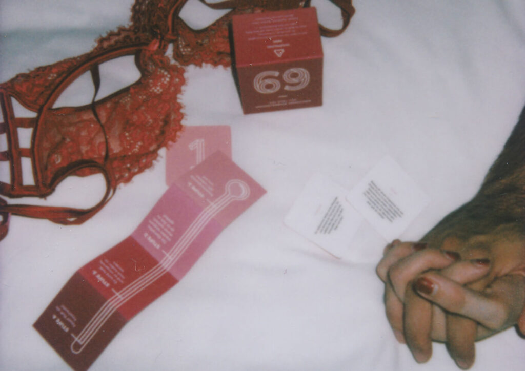 Red lace lingerie and a 69 couples game box with instruction cards and two hands clasped on a white bedsheet
