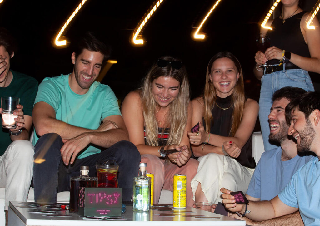 Six young adults laughing and playing a card game, "Tipsy", around a table with drinks at night