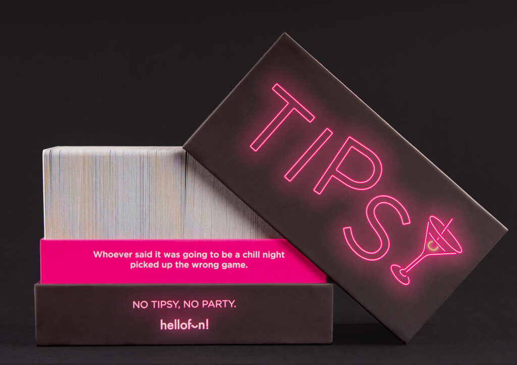 Tipsy card game box, dark with neon pink title &amp; martini glass, open showing a stack of white cards