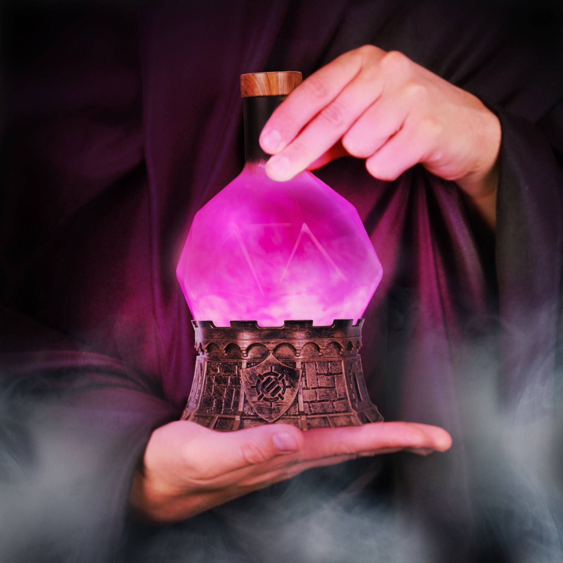 A hand in dark robes holds a glowing pink faceted crystal in a castle-like base, amidst swirling mist