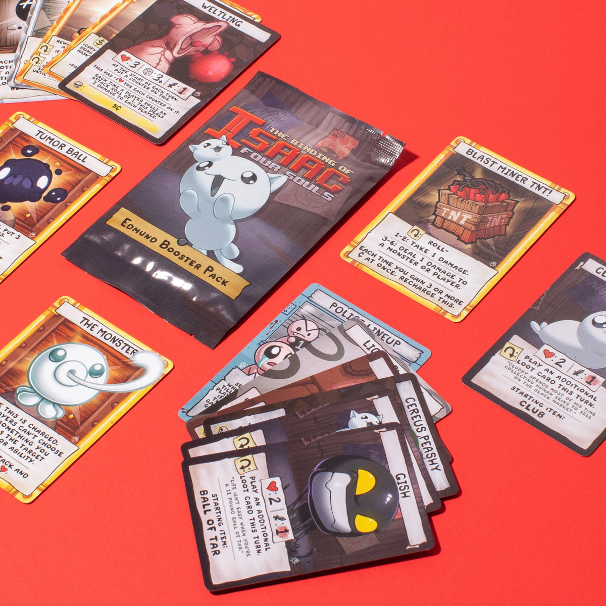 The Binding of Isaac Four Souls Edmund Booster Pack and scattered game cards displayed on a vibrant red surface