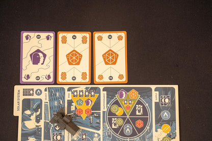 A gray spaceship token rests on a board game showing "Solar System" and "Risk" sections, with three colorful cards nearby