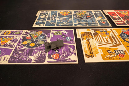 A grey futuristic miniature rests on a purple board game tile. Other blue, red, and yellow game boards are visible on a black table